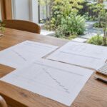 庭の窓辺の机で、外構工事の書類を計算機と並べて照合する場面