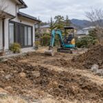 冬の住宅庭で掘削した土、分けた石、停止中の小型重機がある外構工事のイメージ
